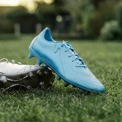 Una coppia di scarpe da calcio, una blu e una bianca, posate sull'erba verde con alberi e un cielo al tramonto sullo sfondo.