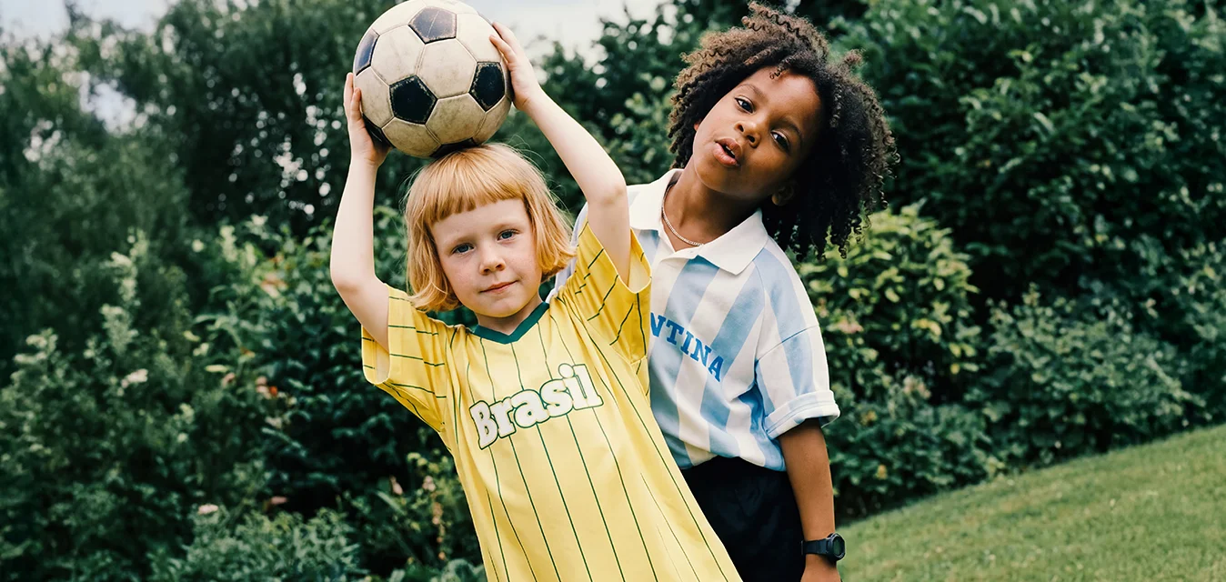 Due bambini in piedi sull'erba, uno con una palla da calcio sopra la testa, indossano le maglie delle squadre di calcio di Brasile e Argentina.