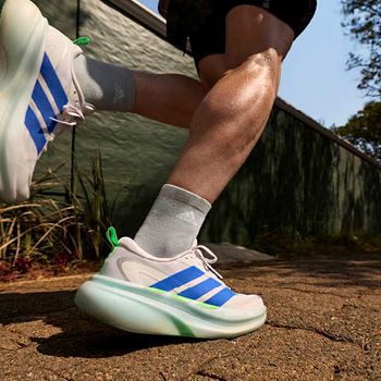 Close-up of runner's legs and white Adidas trainers with blue stripes and green heel tab on sunlit pavement