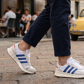 Close-up of person walking on cobbled street wearing white trainers with blue stripes and navy trousers, blurred café and yellow car