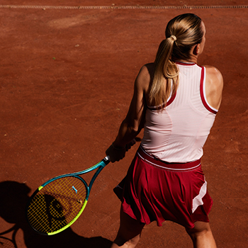 Female tennis player with blonde ponytail hitting a backhand on red clay court, wearing white top and red skirt and holding a racket