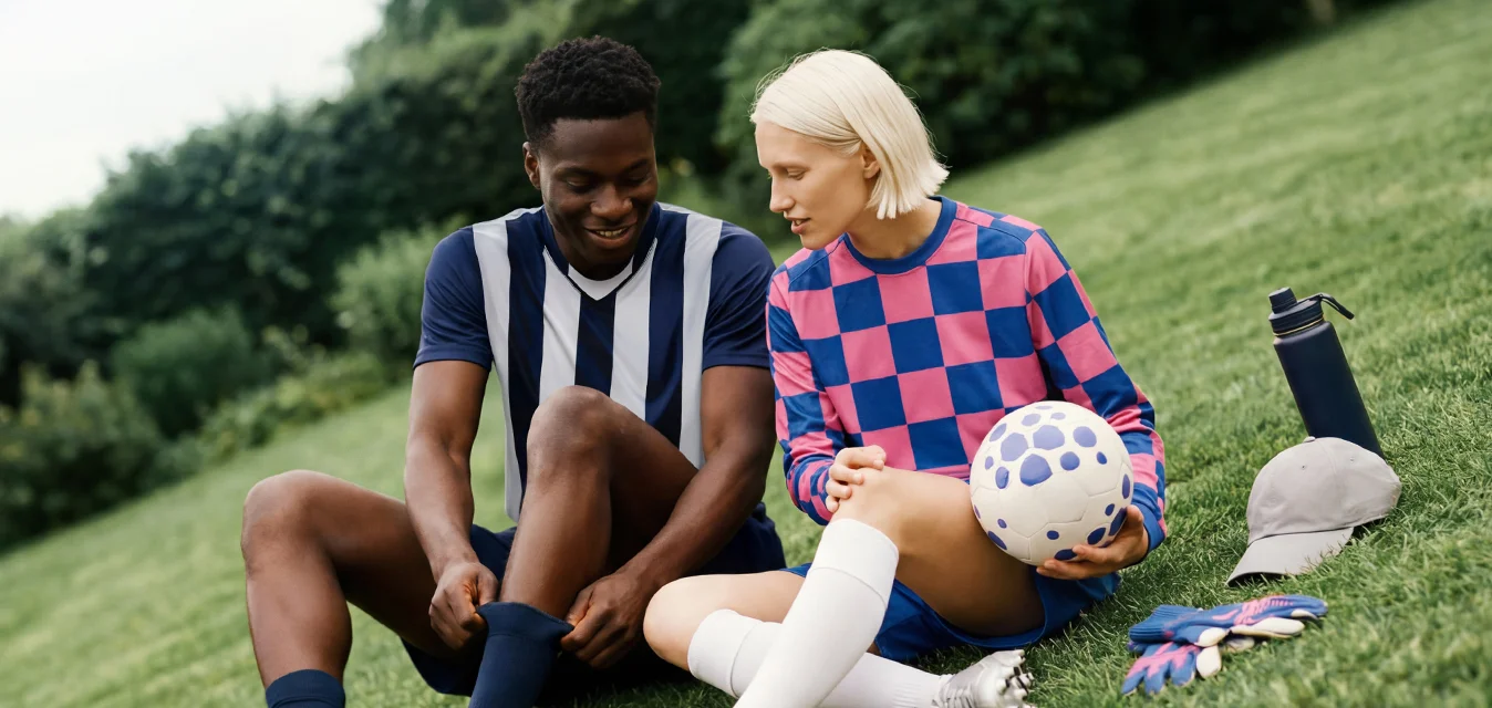 Due giovani in divisa da calcio siedono sull'erba, uno tiene un pallone da calcio, riposandosi con bottiglie d'acqua e cappelli nei dintorni in un parco.