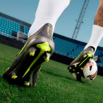 Close-up of football boots with lime-green studs striking a white football on grass at a stadium