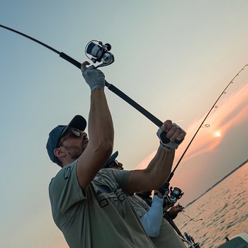 Anglers on a boat casting spinning rods at sunset over calm water, man in cap and gloves reeling in line