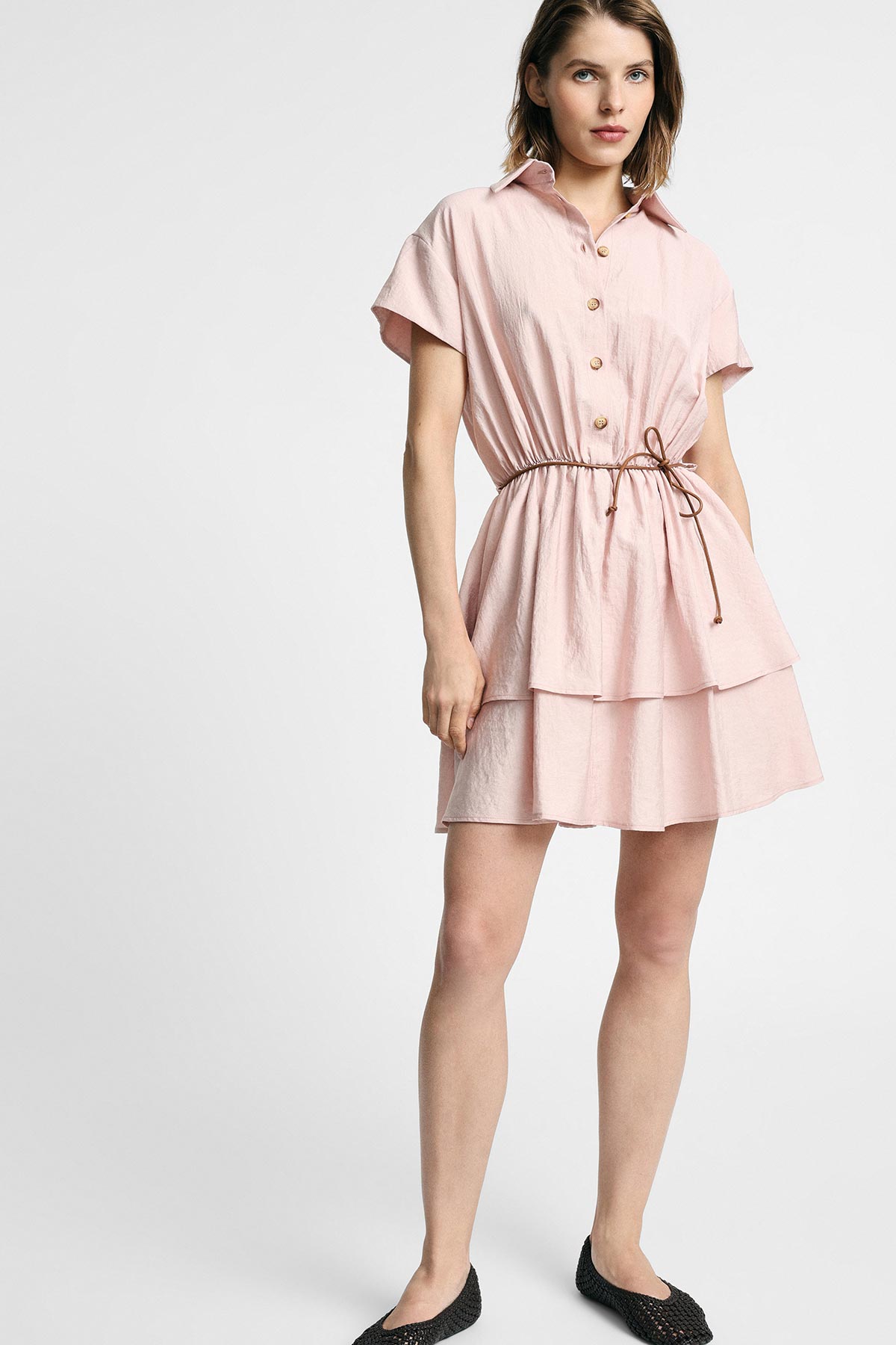 Female model wearing short-sleeve pale pink tiered shirt dress with drawstring waist and black ballet flats on white background