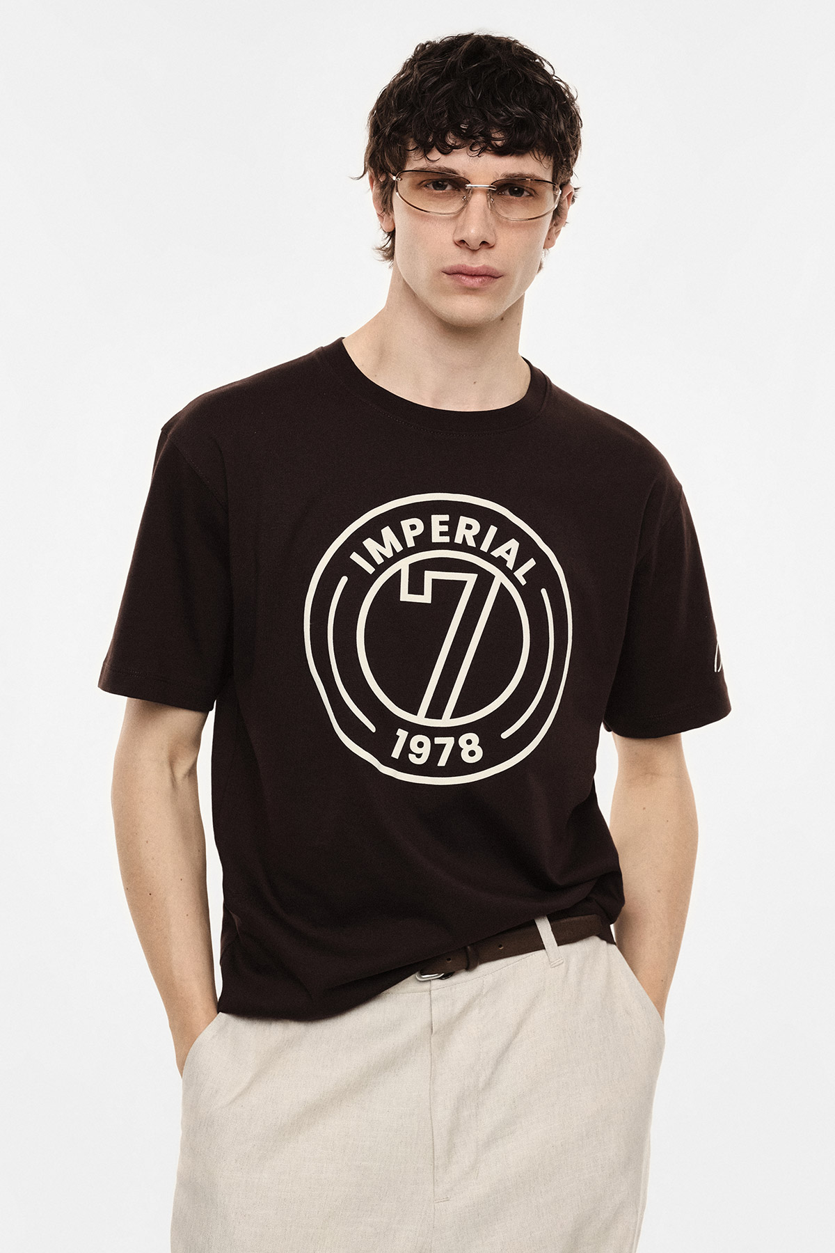 Young man wearing black T‑shirt with large 'Imperial 7 1978' circular logo, beige trousers and sunglasses on a white background
