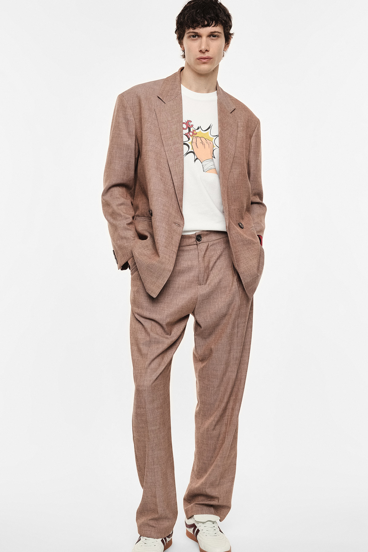 Male model in a relaxed brown tailored suit with a graphic white T‑shirt and white trainers, hands in pockets