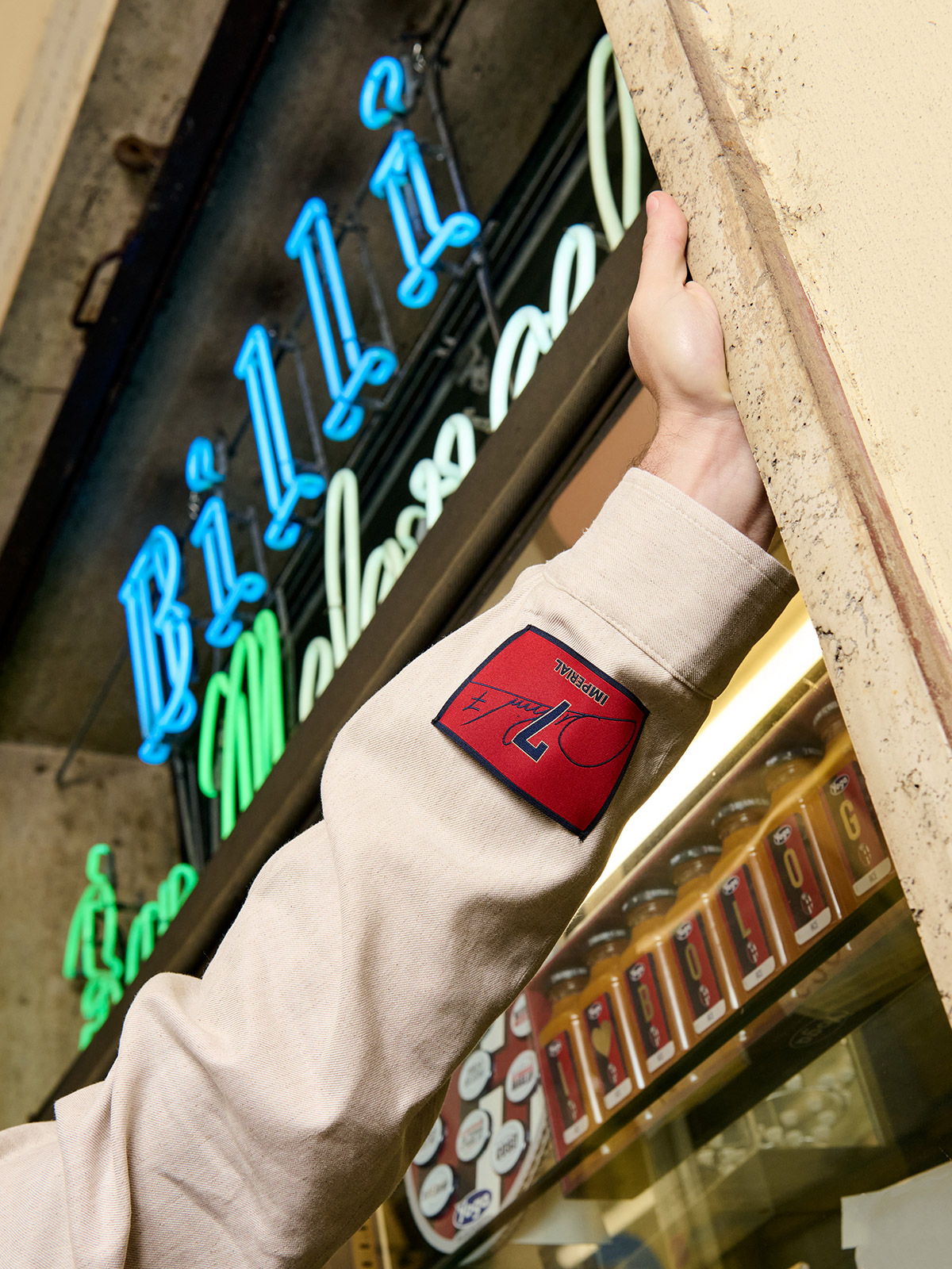 Arm in beige jacket with red logo patch reaching through a pizzeria doorway, blue neon "Pizza" sign and rows of canned goods visible