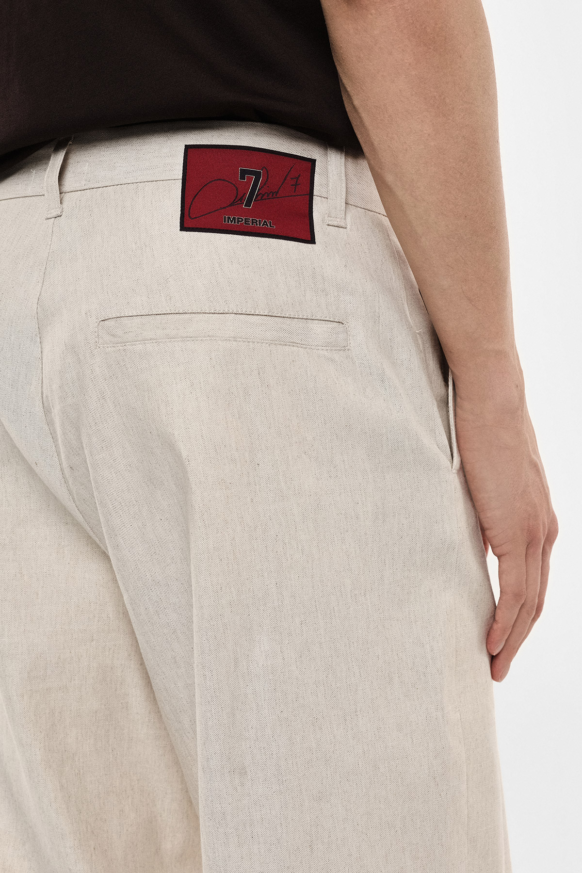 Rear view of beige trousers showing a back welt pocket and a red square brand patch reading 'Imperial' near the waistband