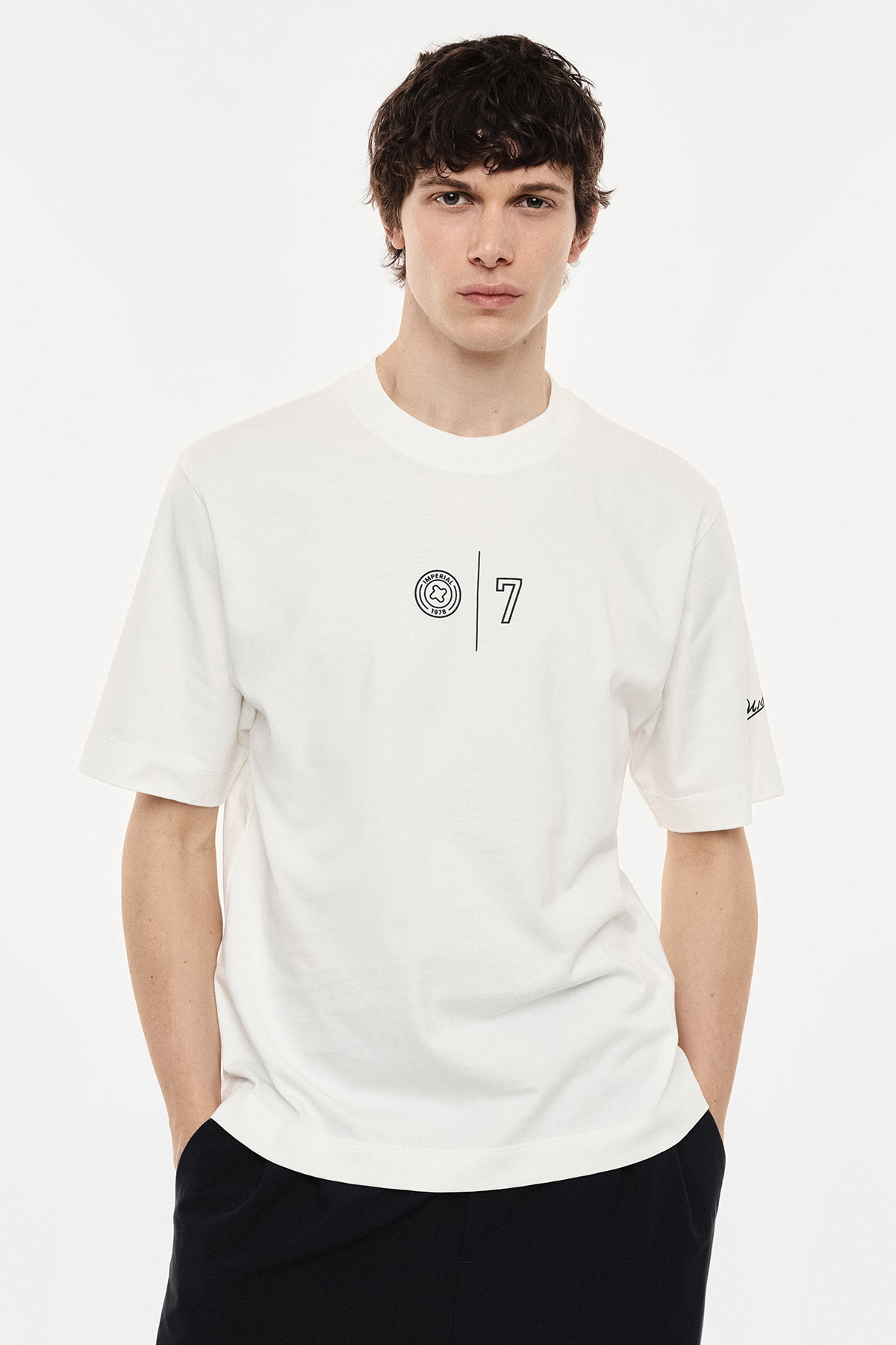 Male model wearing white logo T-shirt with small centred '7' emblem, standing with hands in pockets against plain white background