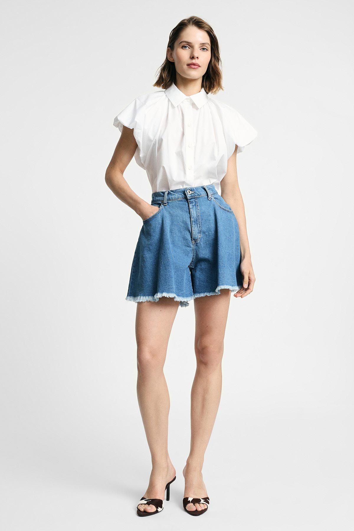 Female model wearing white collared blouse tucked into high-waisted frayed denim shorts and black heeled sandals