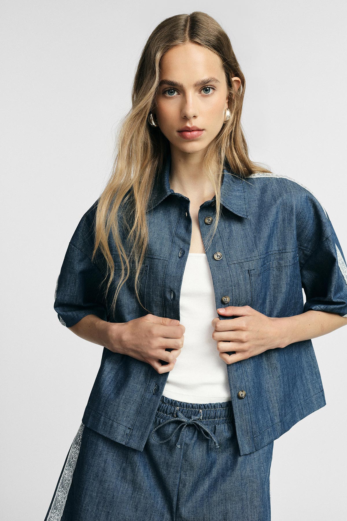 Young woman modelling cropped denim shirt and matching drawstring trousers over a white ribbed tank top on a neutral background