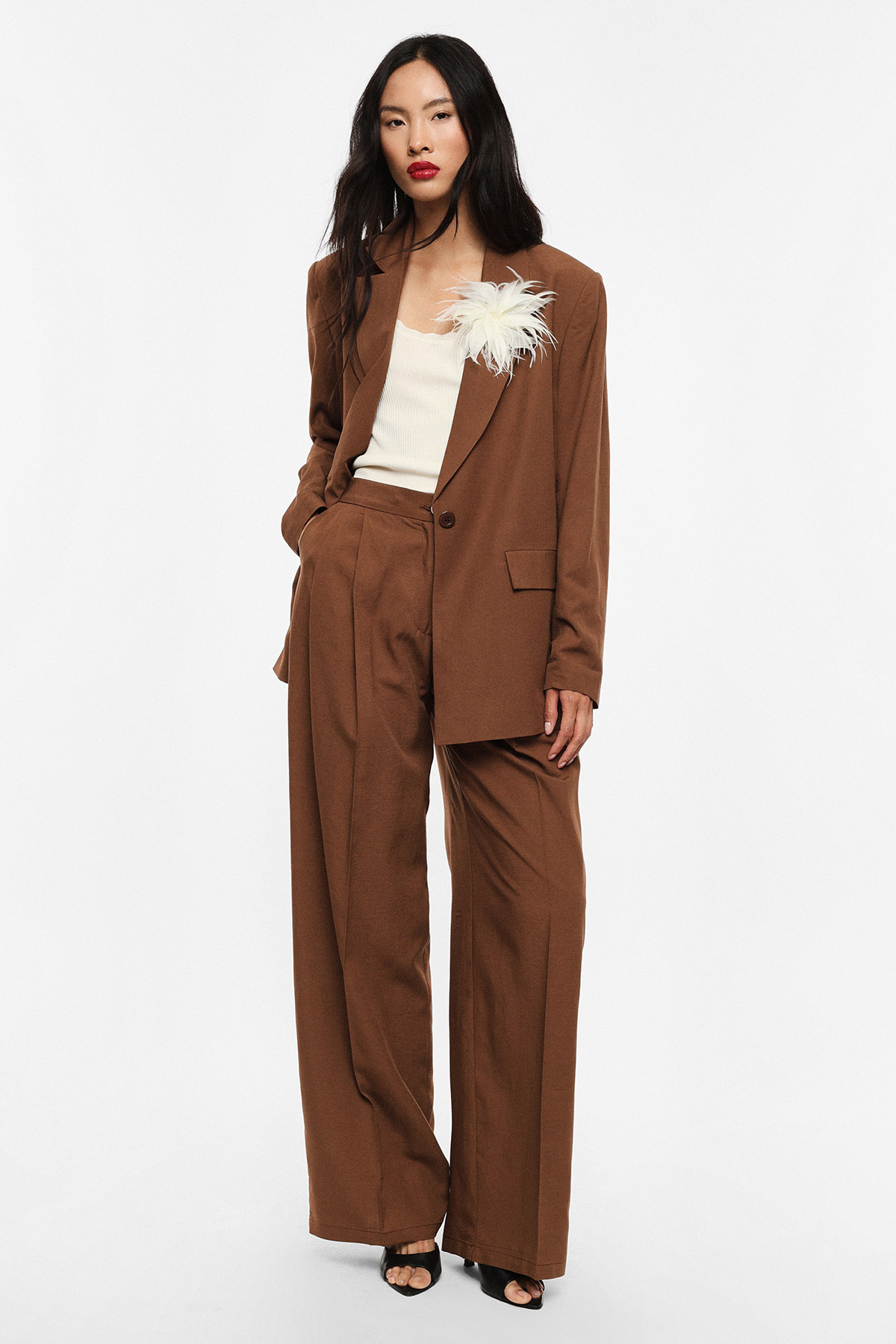Model in brown oversized suit with wide-leg trousers, cream camisole and white feather brooch on a white background