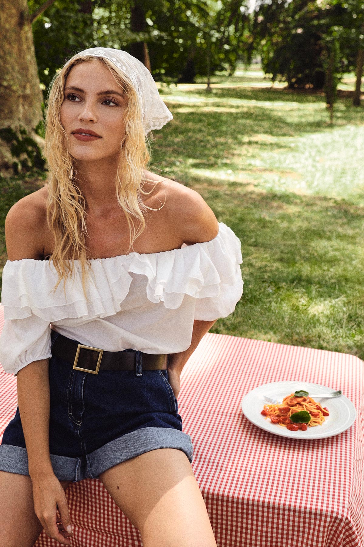 Young woman in white bardot top and denim shorts sitting on a red-check picnic blanket with a plate of pasta in a sunny park