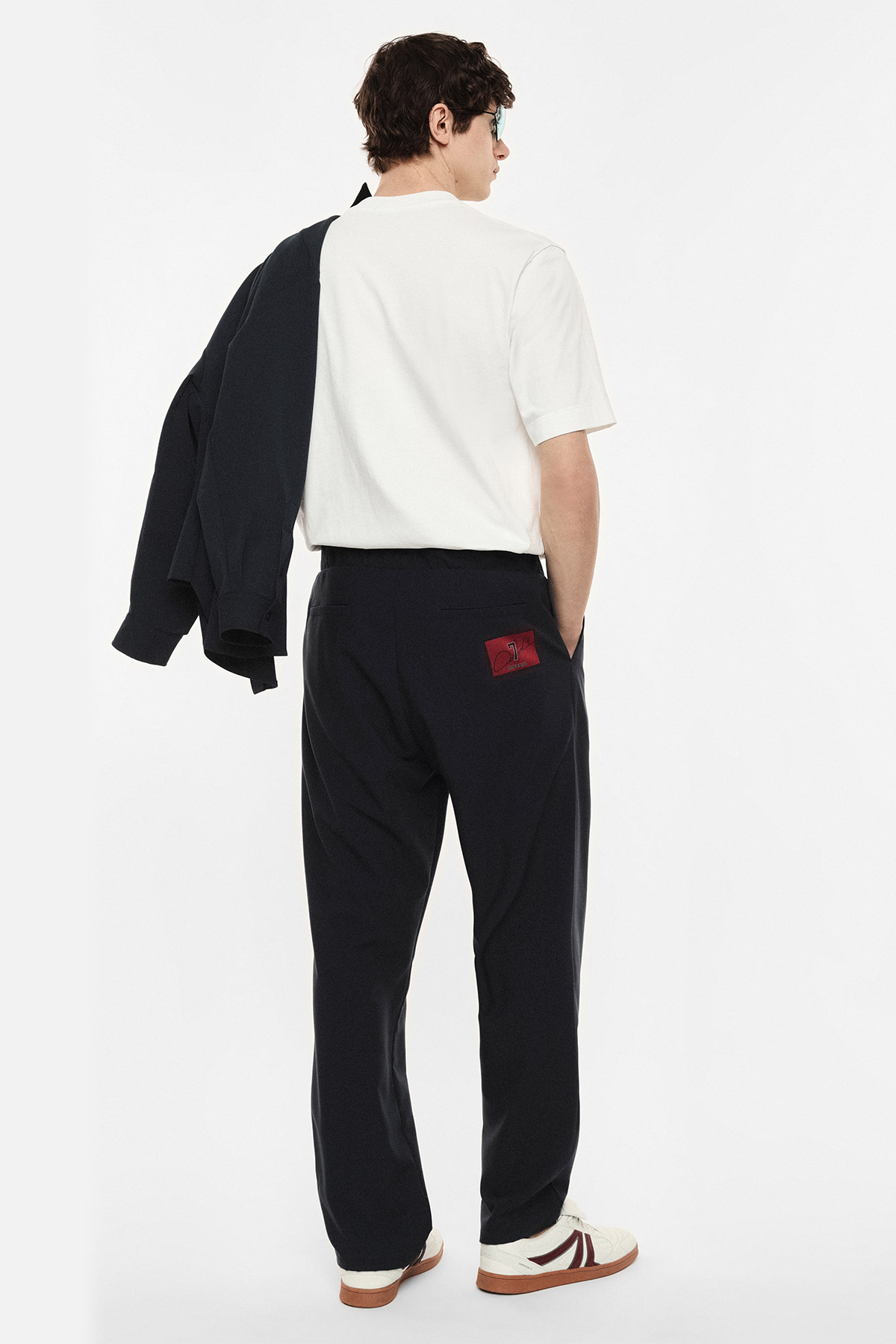 Man seen from behind wearing a white T-shirt, navy trousers with a red patch, navy jacket draped over one shoulder and white trainers