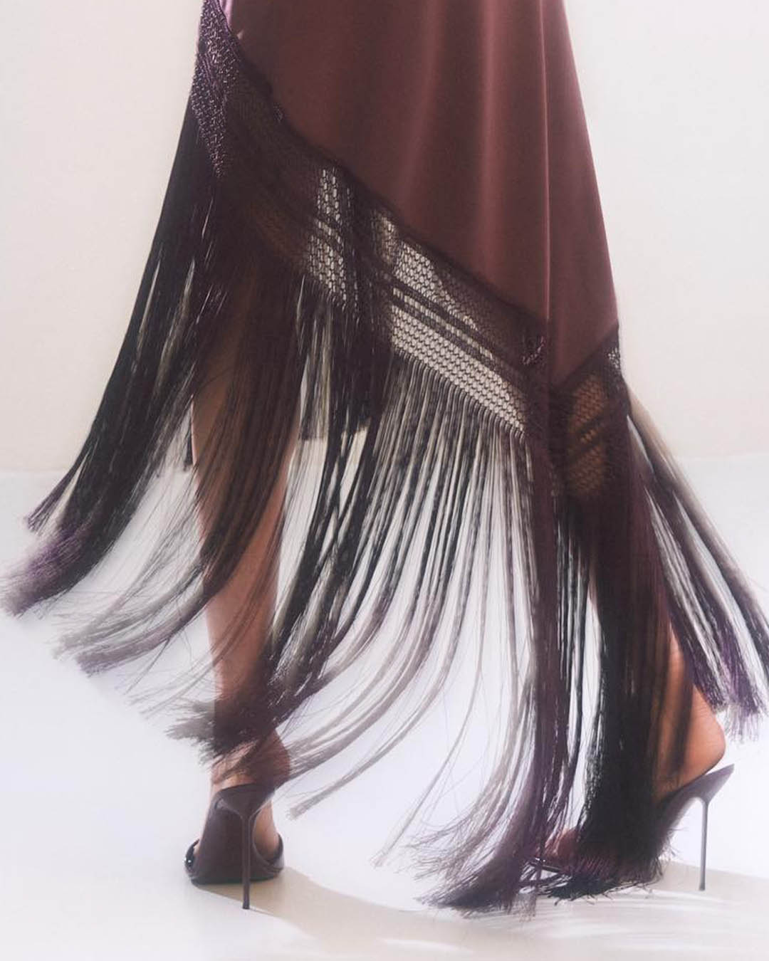 Legs in motion wearing a burgundy fringe dress with long tassels and black stiletto heels on a white background