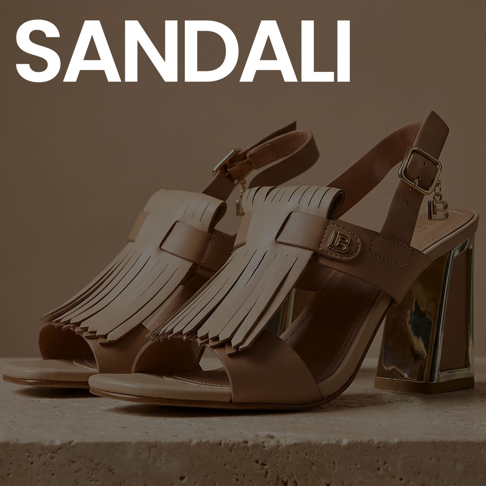 Pair of tan leather high-heeled sandals with fringe detail, metallic block heels and ankle buckle strap on beige backdrop
