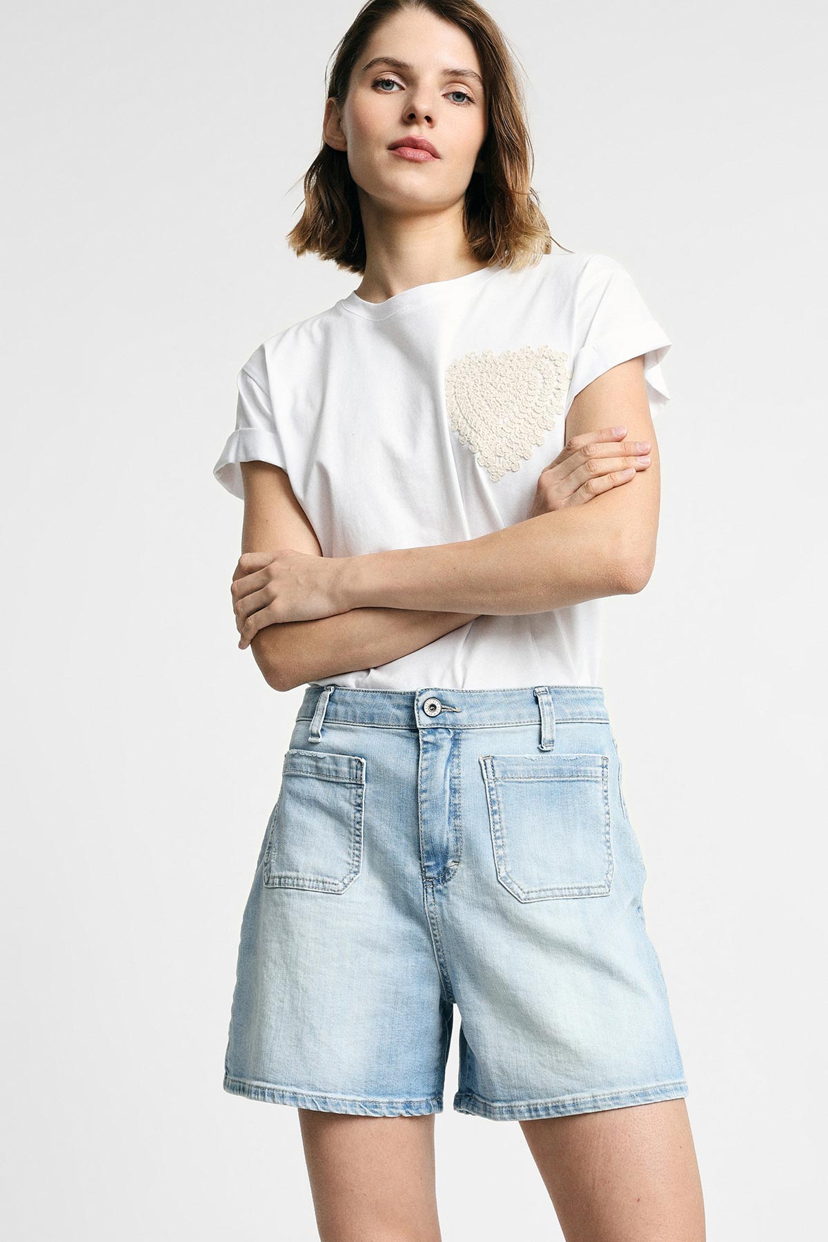 Young woman standing with arms crossed wearing white T‑shirt with textured heart patch and light denim shorts on plain white background