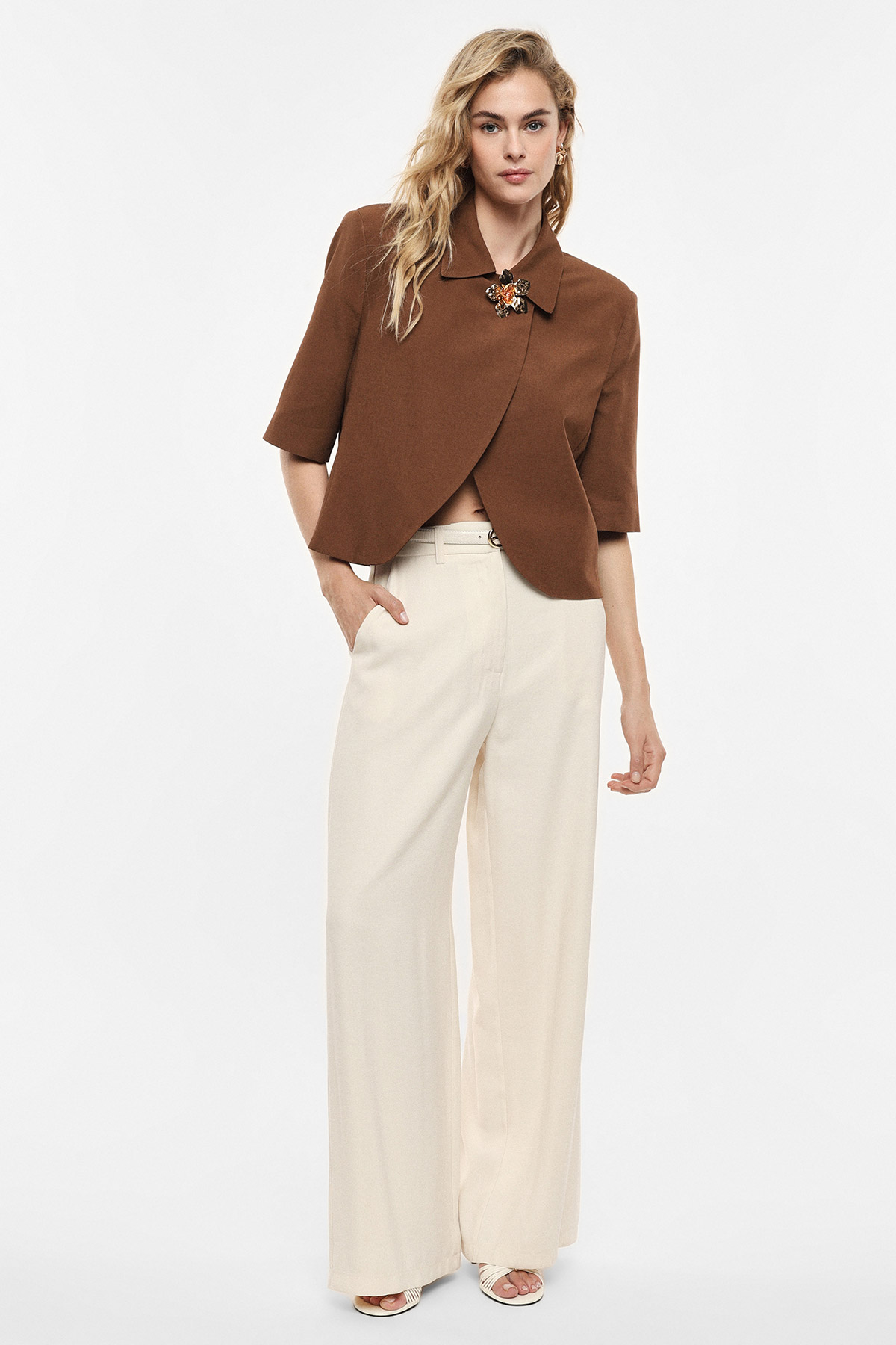 Woman wearing a brown cropped jacket with brooch and cream wide‑leg trousers on a white background