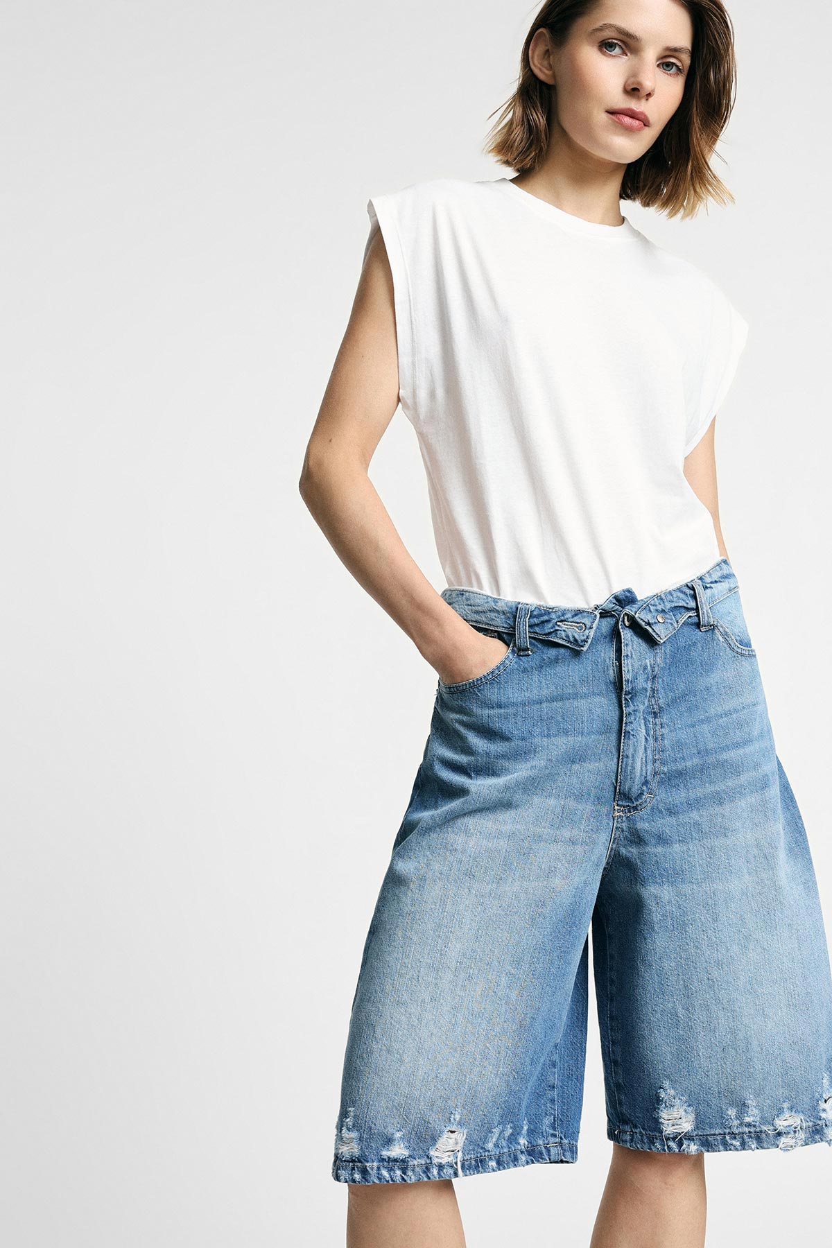 Woman in white sleeveless top and knee-length distressed denim shorts, hands in pockets against plain light background