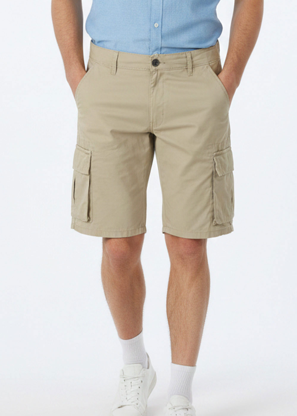Man wearing beige cargo shorts with side flap pockets, light blue tucked shirt, white socks and trainers, hands in pockets