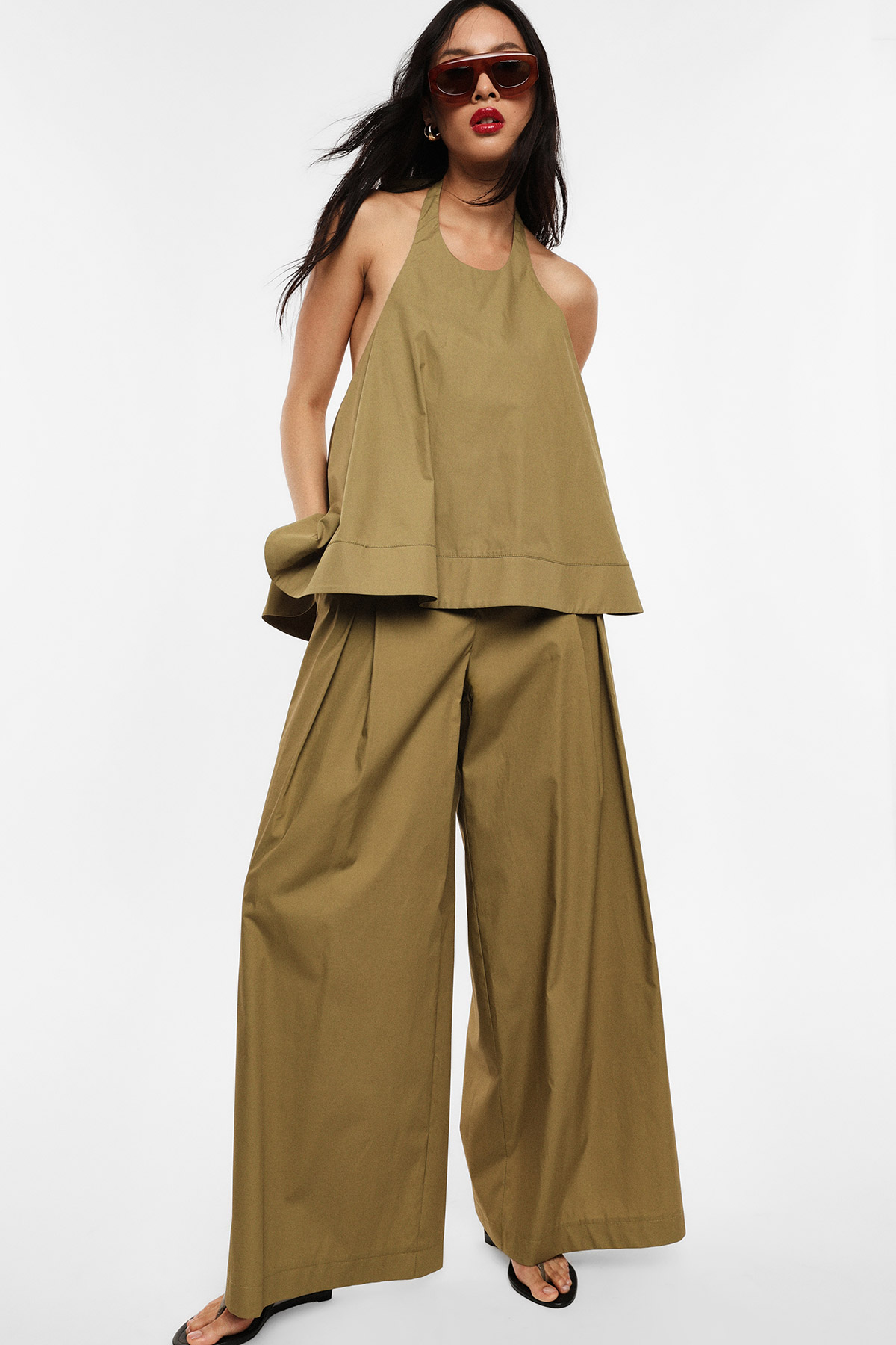 Model in olive‑green halter top and wide‑leg trousers, wearing sunglasses and sandals against a white background
