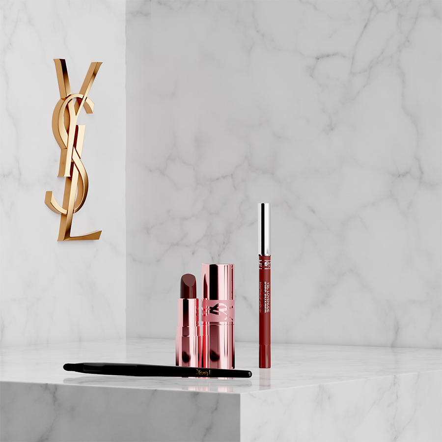 Un rossetto YSL in un astuccio metallizzato oro rosa, una matita labbra e un pennello nero esposti su un piedistallo di marmo bianco accanto a un logo YSL dorato.