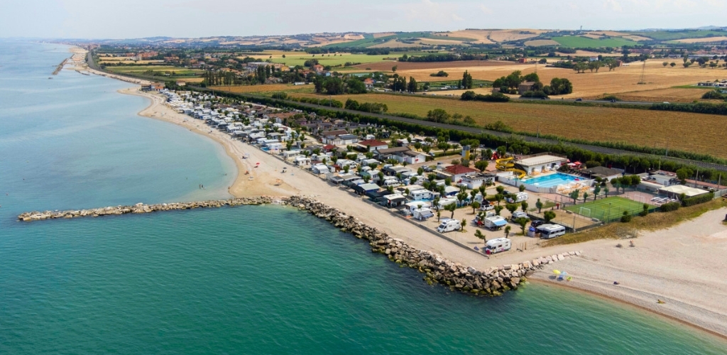 Aerial view of a coastal caravan park and campsite on a sandy beach with turquoise sea, outdoor pool, tennis courts and rolling farmland