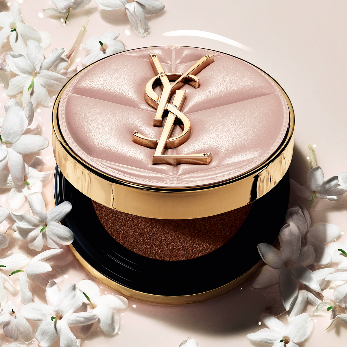 Un fondotinta cushion YSL in un astuccio trapuntato rosa e oro, circondato da delicati fiori di gelsomino bianchi.