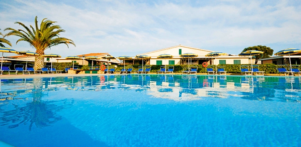 Resort outdoor swimming pool with clear blue water, palm tree, sun loungers and parasols in front of white holiday villas