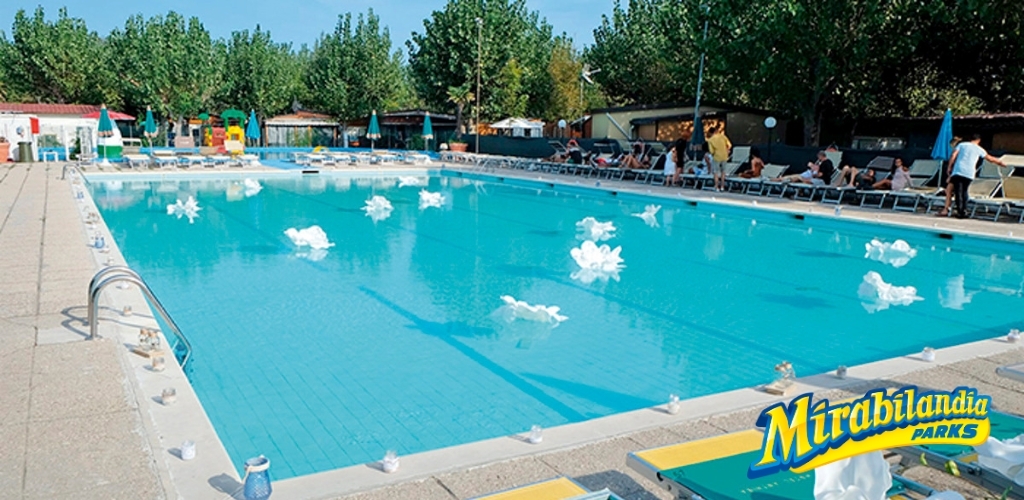 Large outdoor swimming pool with sun loungers and guests relaxing under trees, Mirabilandia Parks logo visible