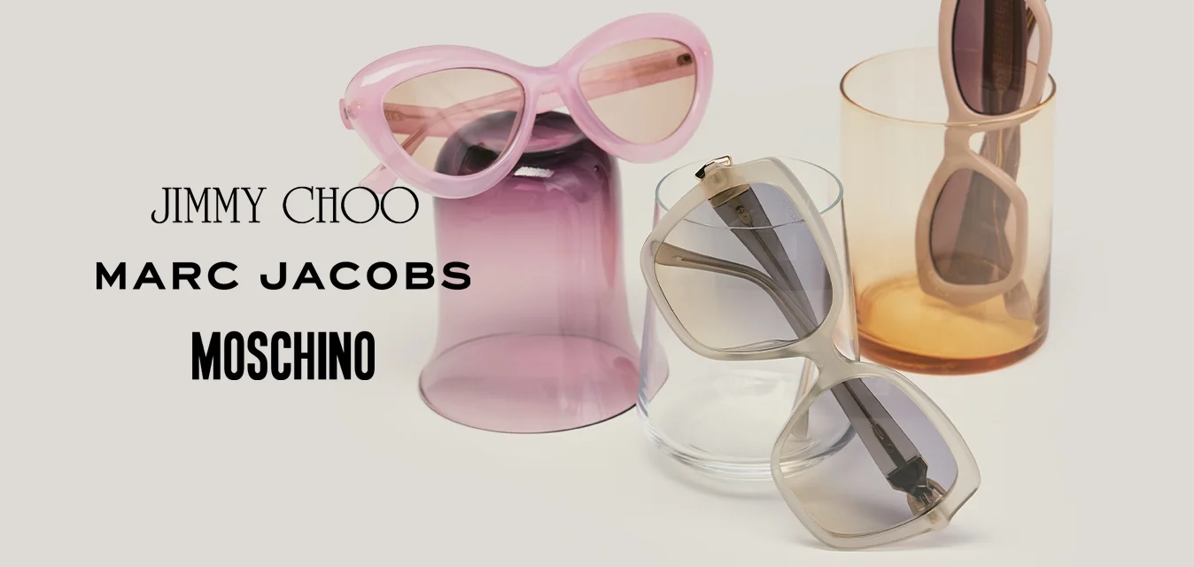 Tre paia di occhiali da sole in rosa, grigio e beige, esposti su e intorno a bicchieri di vetro traslucido viola, trasparente e ambra.