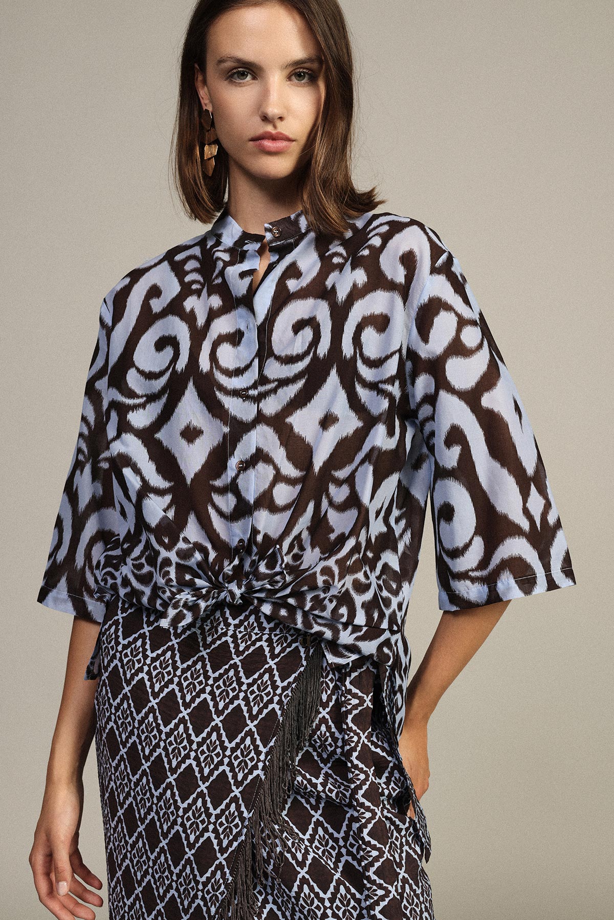 Woman wearing blue-brown patterned blouse knotted at the waist with a geometric-print skirt and gold earring