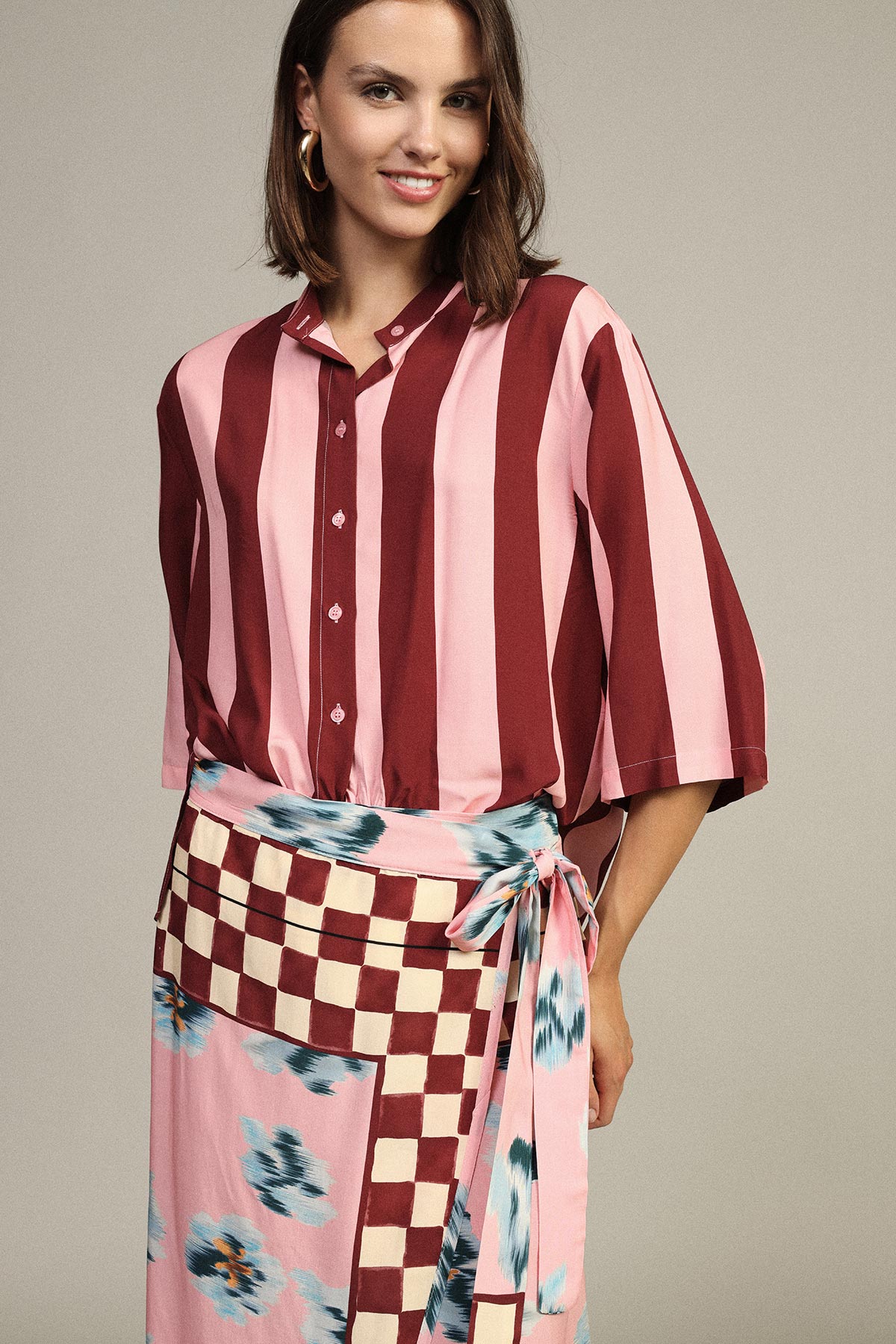 Woman in pink and burgundy striped blouse and wrap skirt with checkerboard and blue floral print, wearing gold hoop earring