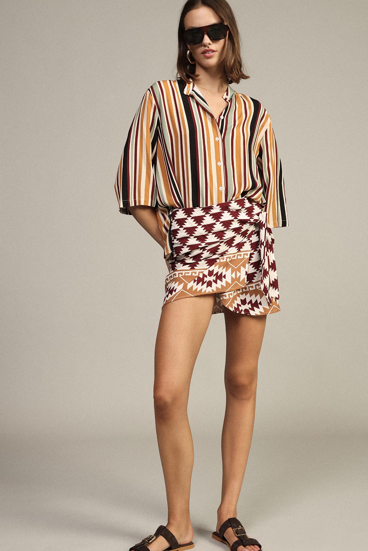 Woman wearing multicolour vertical striped shirt, geometric wrap skirt, black sunglasses and sandals in a casual summer fashion pose