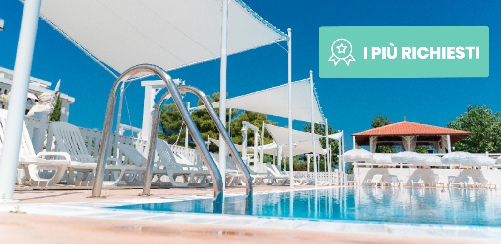 Resort swimming pool with stainless-steel ladder, rows of white sunloungers and shaded canopies beneath a clear blue sky