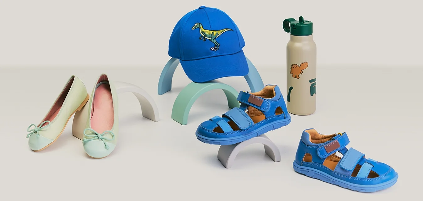 Coppia di ballerine verde pallido da donna, sandali blu per bambini, cappellino blu con dinosauro e borraccia beige con stampe di dinosauri su superficie bianca.