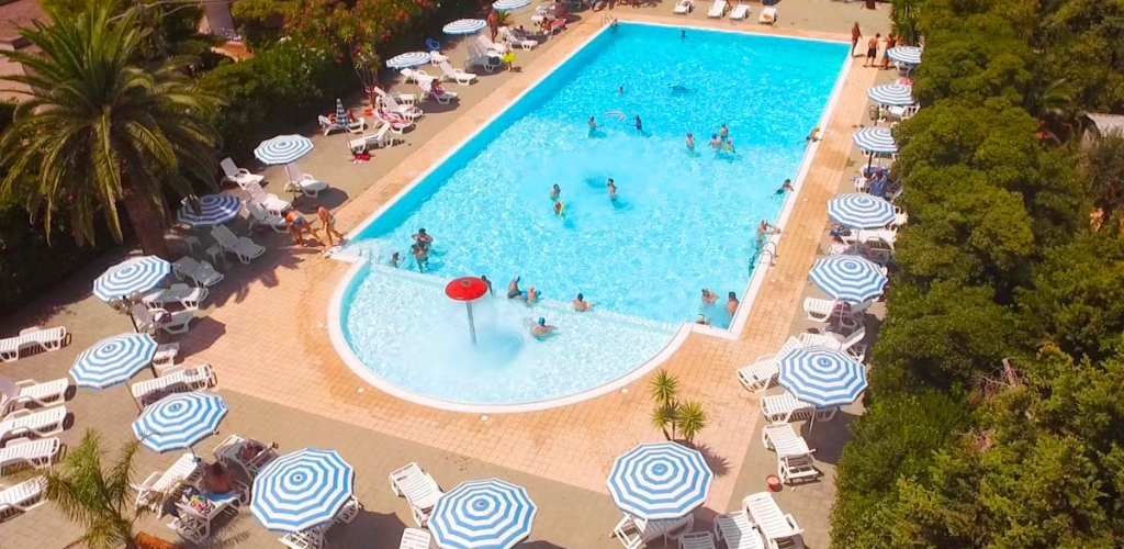 Aerial view of a busy outdoor swimming pool with swimmers, blue-and-white parasols, sunloungers and palm trees on a sunny day