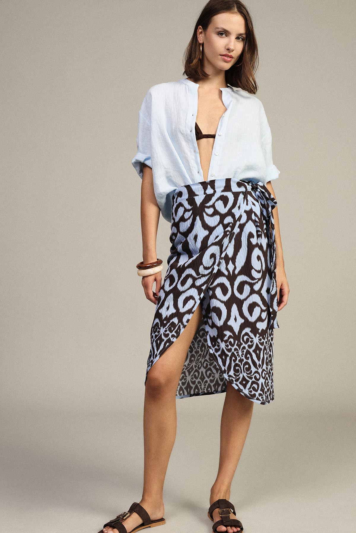 Female model wearing a light blue open linen shirt, black bikini top and patterned blue-brown wrap skirt with side slit and brown sandals