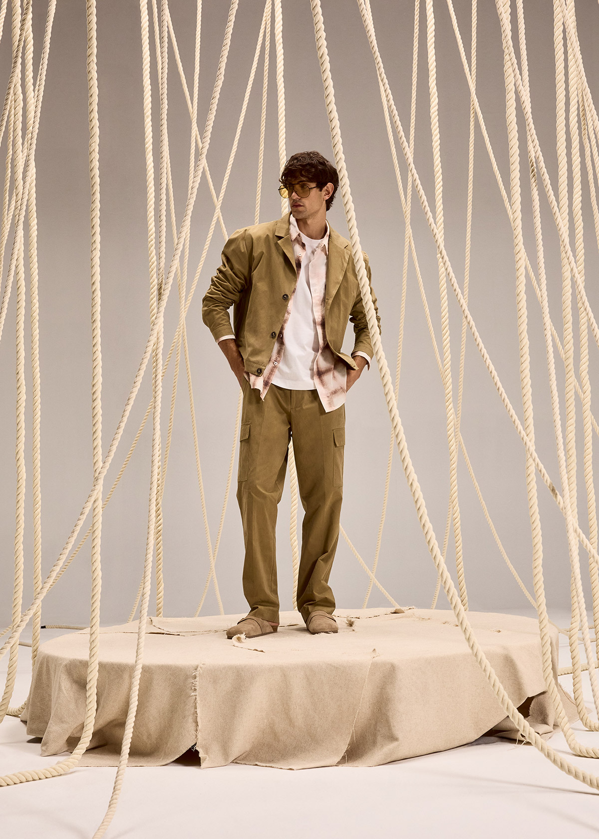 Man in khaki jacket and cargo trousers standing on a fabric-covered platform surrounded by hanging ropes in a minimalist studio