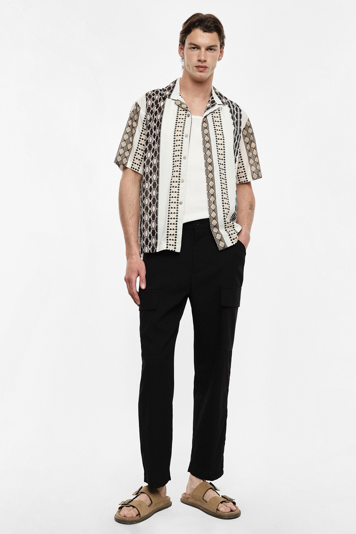Male model wearing patterned short-sleeve shirt over white vest, black cargo trousers and tan sandals