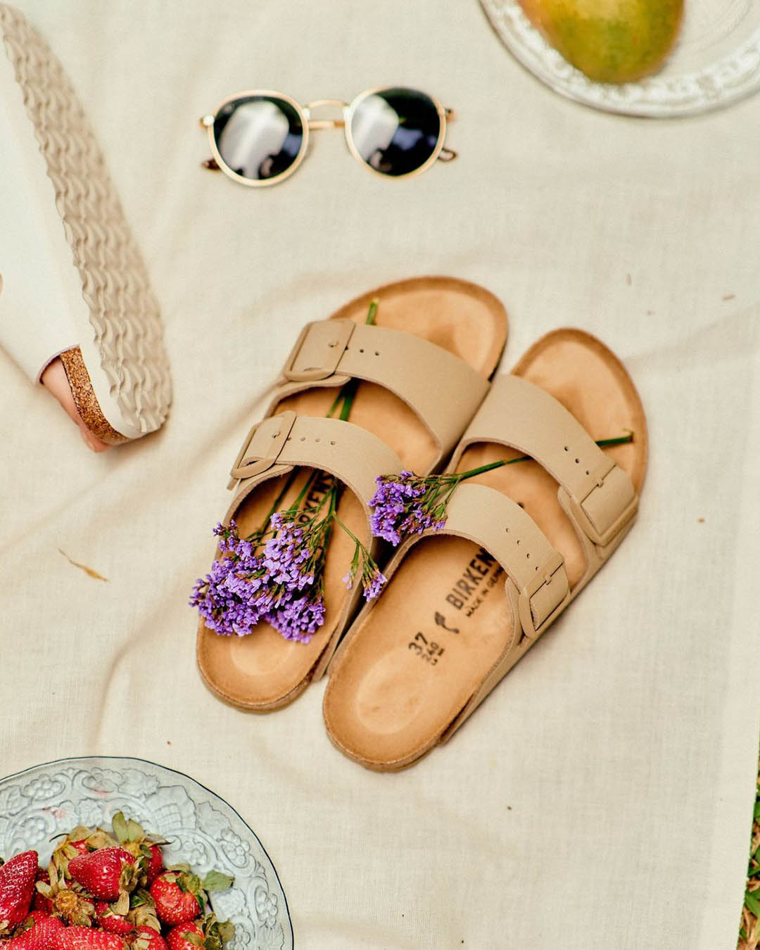 Beige Birkenstock sandals with purple wildflowers tucked inside, round sunglasses and a bowl of strawberries on a picnic blanket