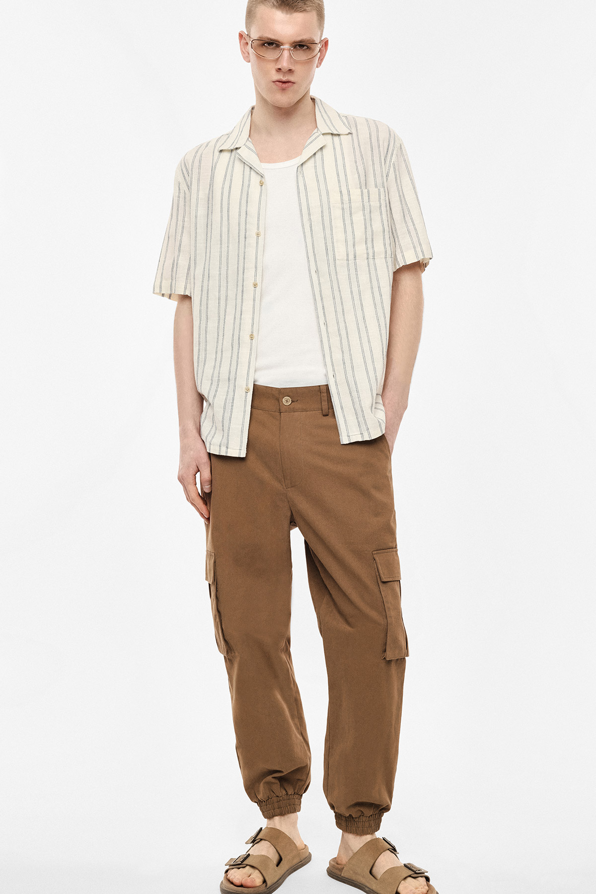 Male model wearing cream striped short-sleeve shirt over white vest, brown cargo jogger trousers and tan sandals, standing with hands in pockets