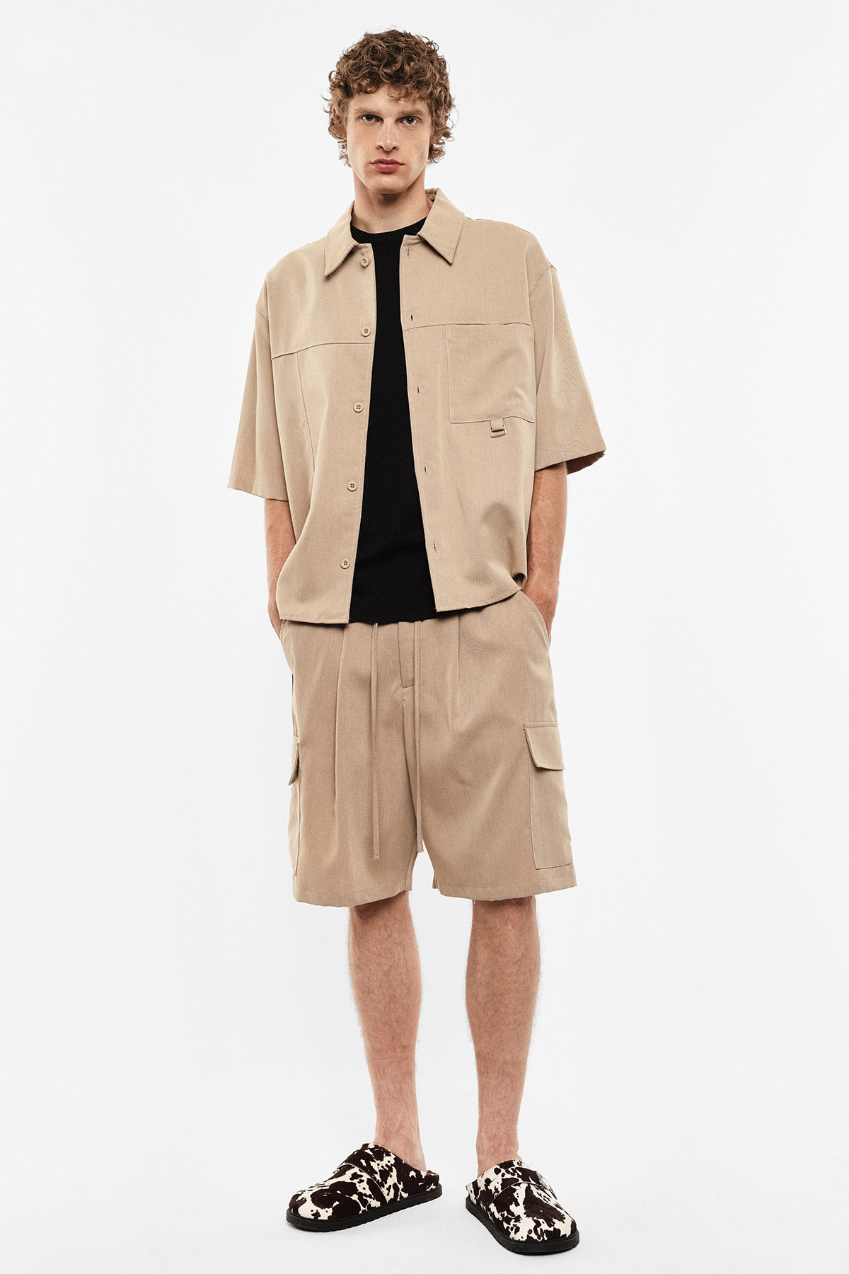 Male model wearing beige short-sleeve overshirt and matching cargo shorts over a black tee, patterned slip-on slippers