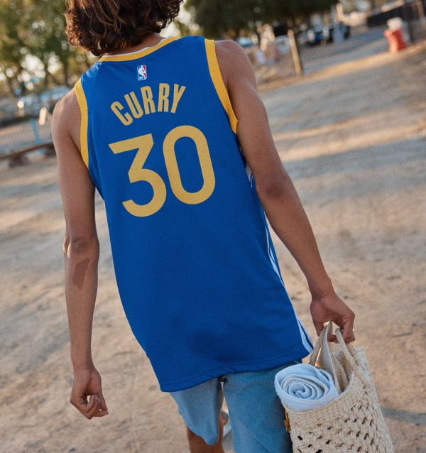 Back view of a person wearing a blue Golden State Warriors Stephen Curry #30 jersey, carrying a woven beach bag outdoors.