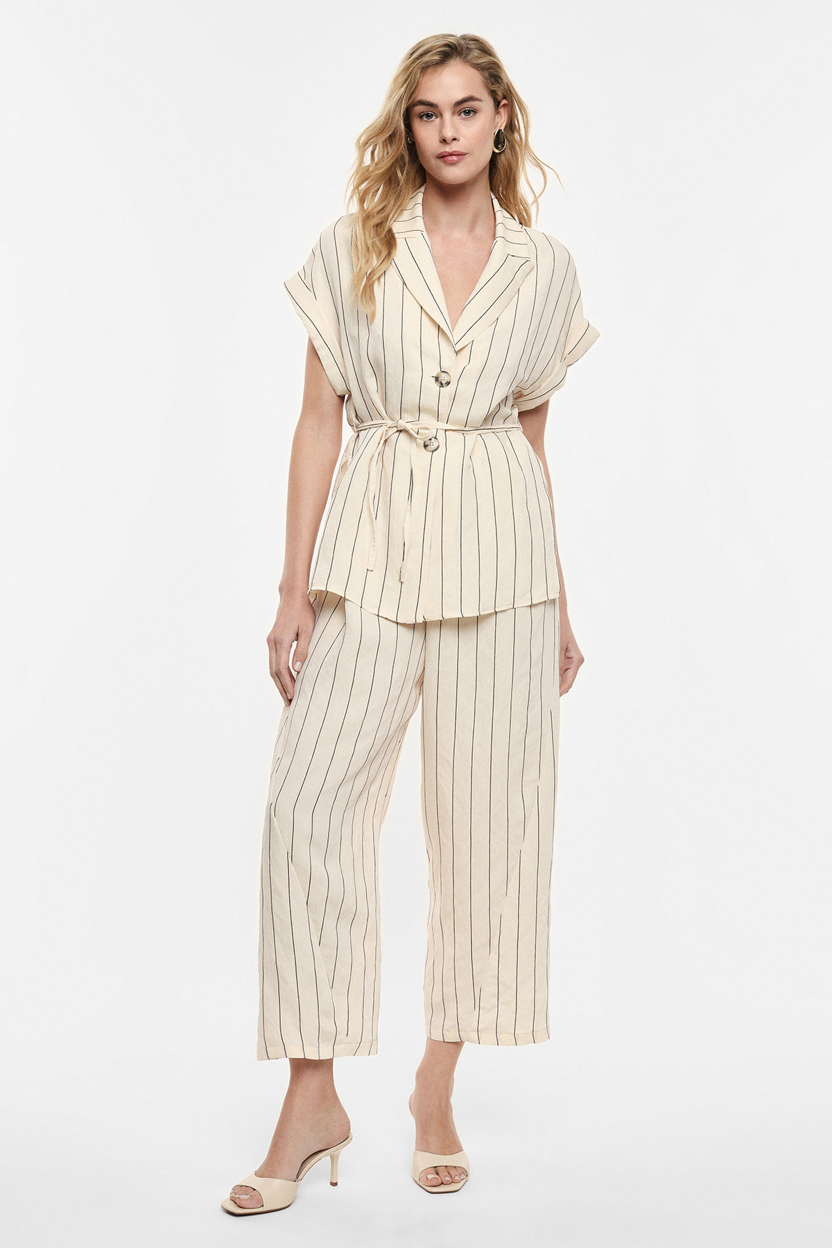 Woman in cream pinstripe belted blazer and matching wide-leg trousers with beige heels on white background