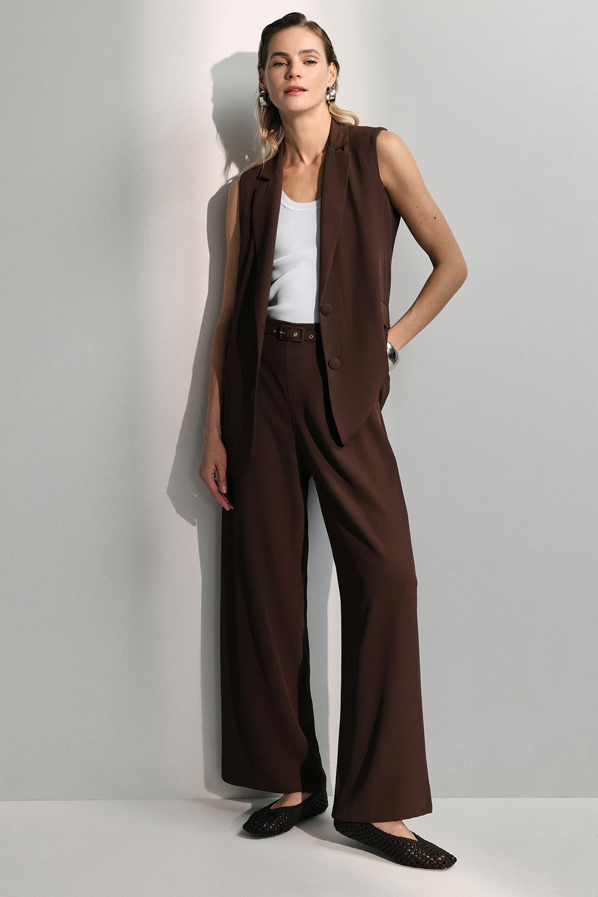 Young woman leaning against wall wearing brown sleeveless blazer and wide-leg trousers, white tank top and black flats
