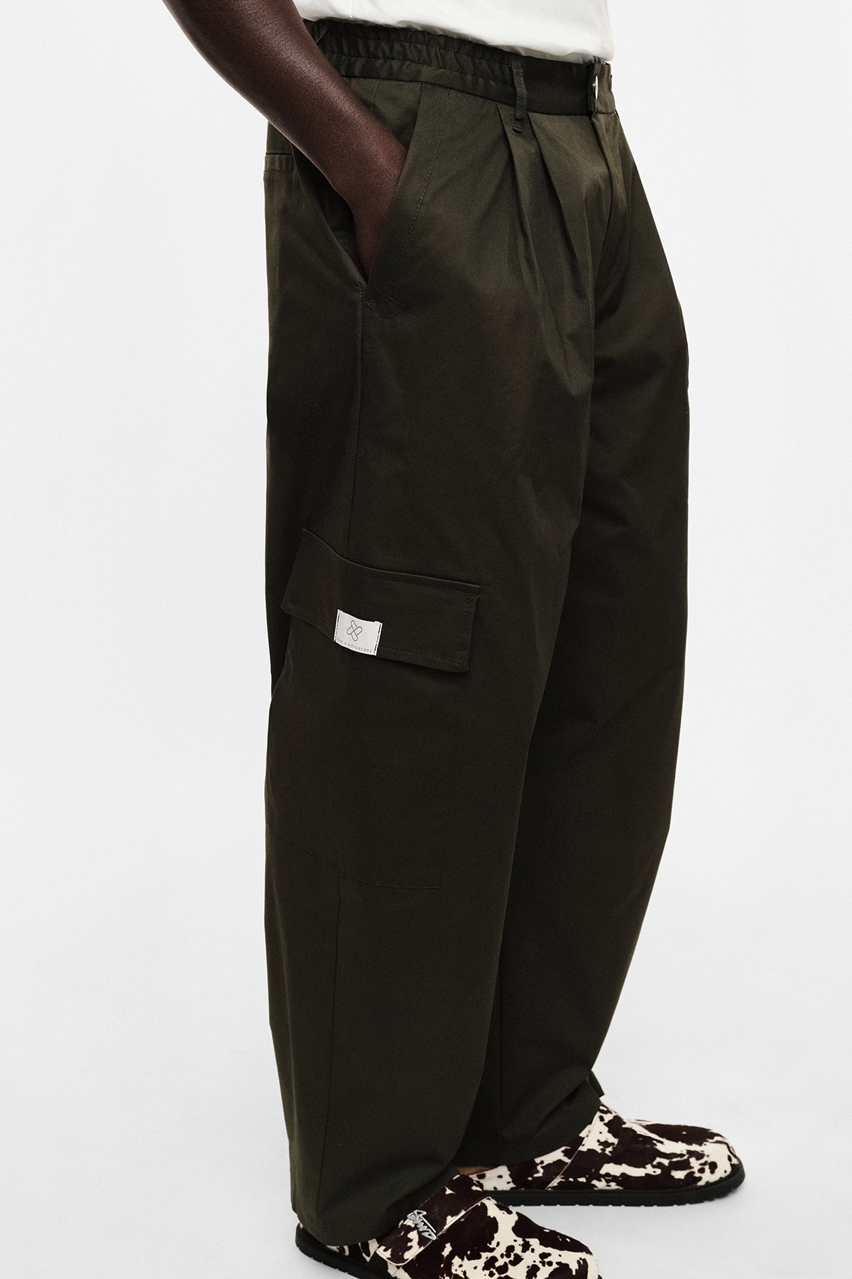 Men's dark olive wide-leg cargo trousers with elastic waistband and side flap pocket, model wearing black-and-white patterned shoes
