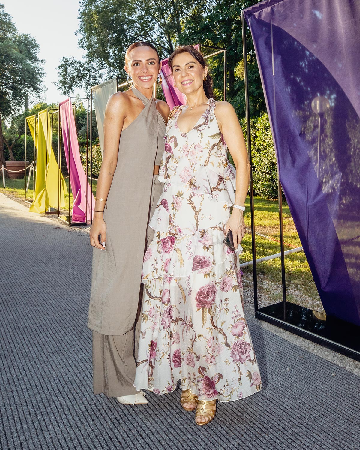 Two women in summer dresses smiling at an outdoor event with colourful fabric panels and trees