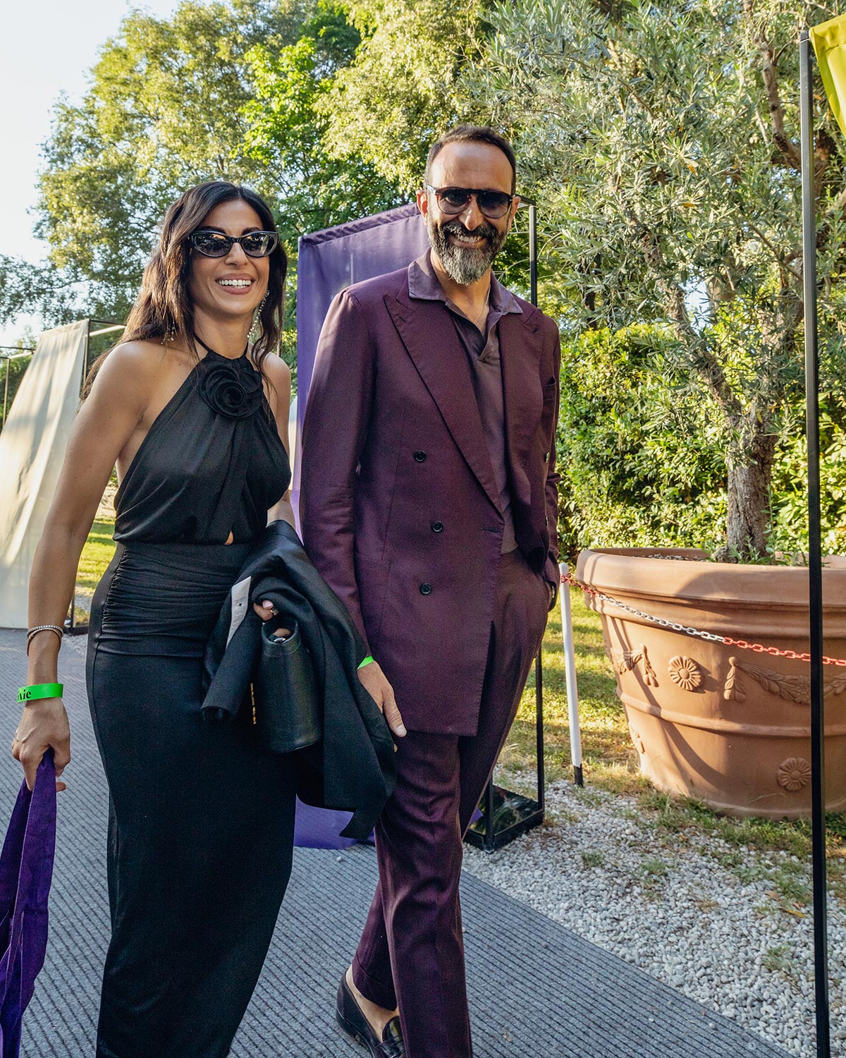 Smiling well-dressed couple at an outdoor event — woman in black dress and sunglasses, man in maroon double-breasted suit and sunglasses
